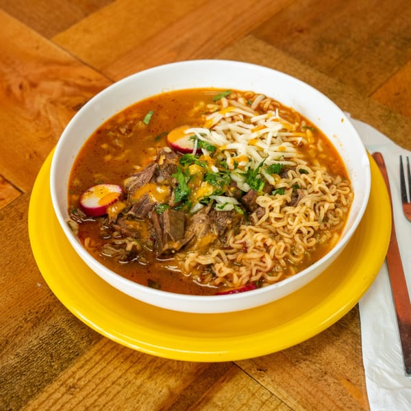 Carnales Taqueria's birria ramen with shredded beef, noodles, cheese, cilantro, and radishes in a white bowl.
