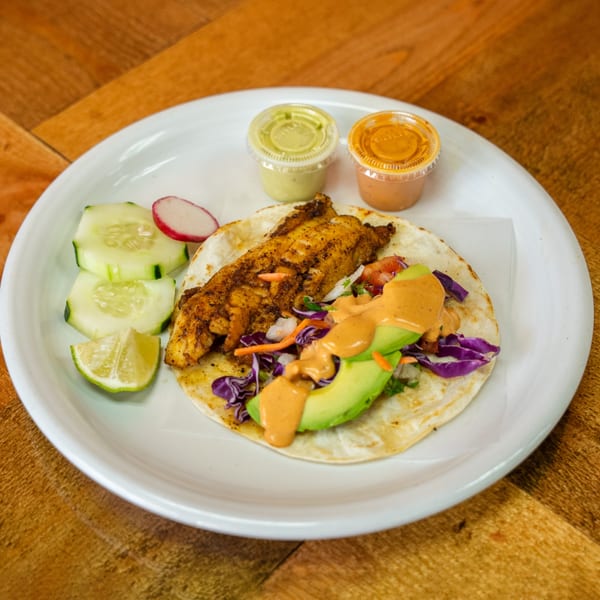 A Carnales Taqueria fish taco with avocado, cabbage, and sauce, served with cucumber, radish, and lime.