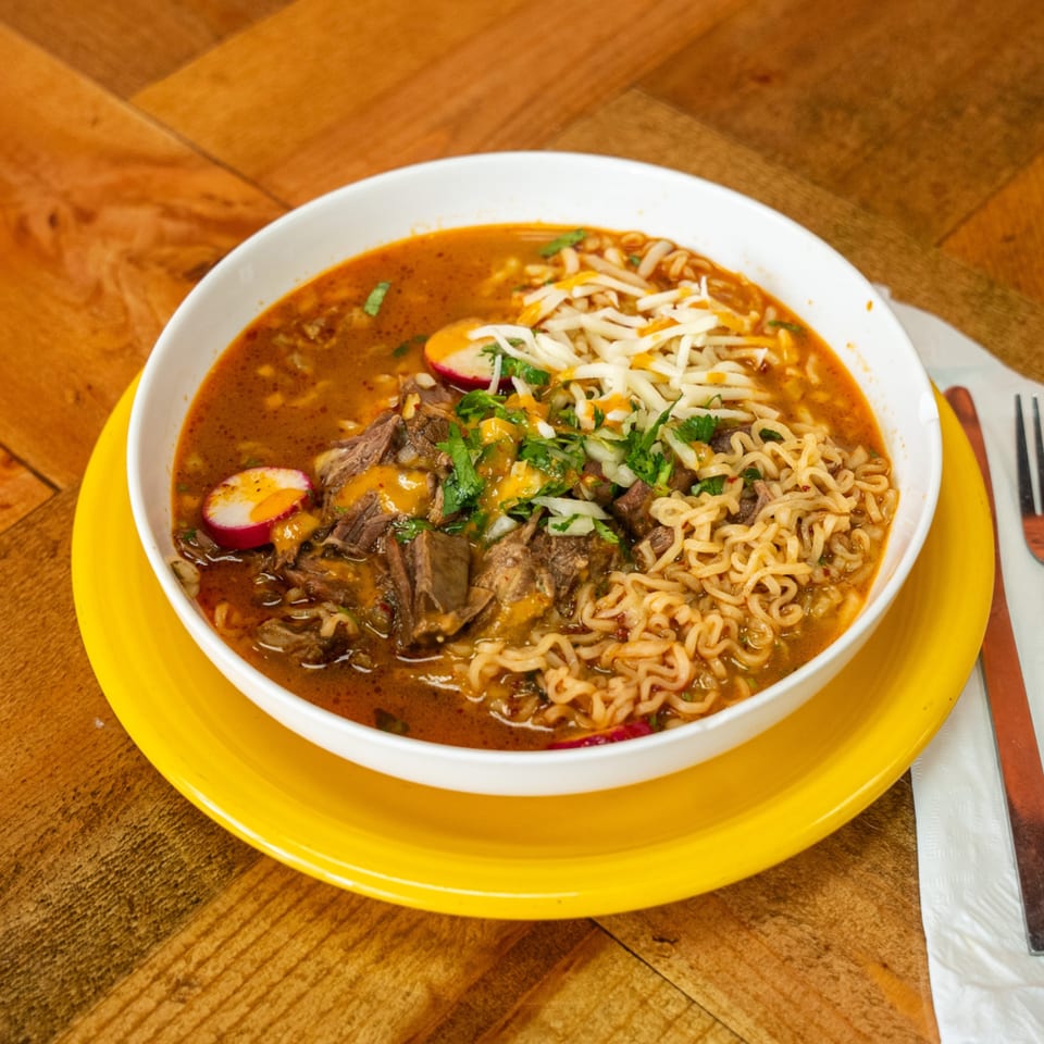 Carnales Taqueria's birria ramen with shredded beef, cheese, cilantro, and radishes in a white bowl.