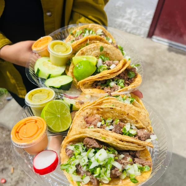 Two plastic trays of Carnales Taqueria tacos with lime, cucumber, radish, and sauces, held by a person.