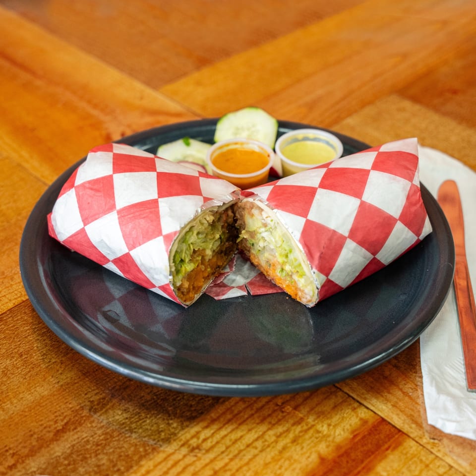 A Carnales Taqueria burrito, cut in half, wrapped in red and white checkered paper, with sauces.