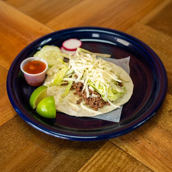 A Carnales Taqueria ground beef taco with lettuce and cheese, salsa, lime, cucumber, and radish.