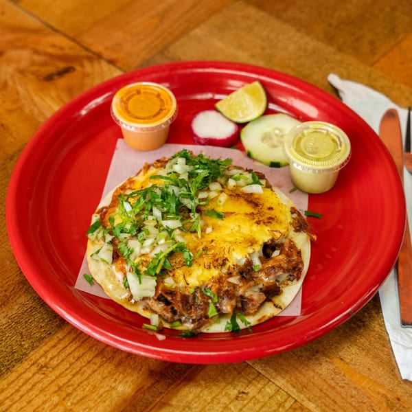 A Carnales Taqueria taco with melted cheese, onions, and cilantro, served with sides on a red plate.