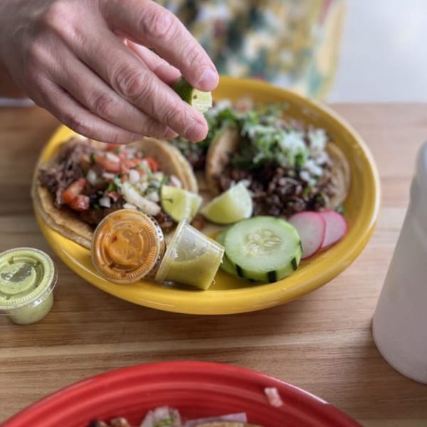 Hand squeezing lime over Carnales Taqueria tacos with sauces, cucumber, and radish on a yellow plate.