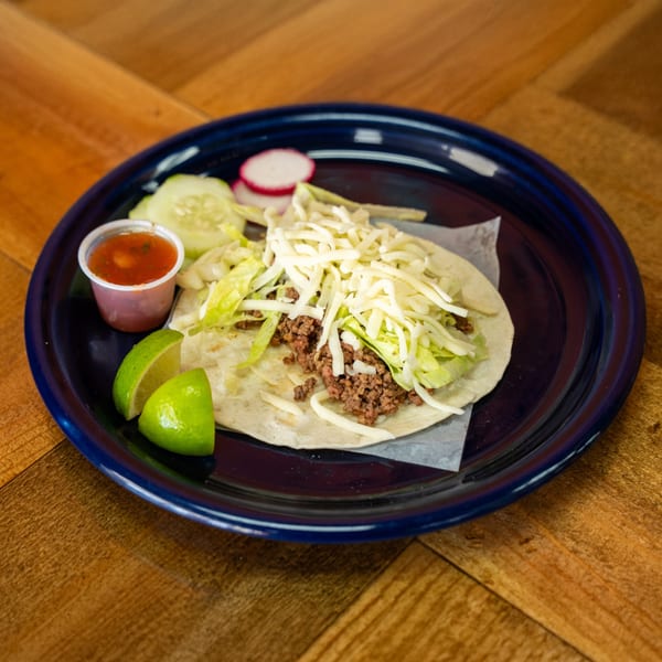 A Carnales Taqueria ground beef taco with shredded cheese and lettuce, served with salsa, lime, and cucumber.