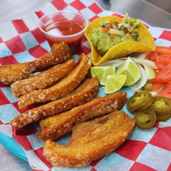 Crispy chicharrones, guacamole taco, salsa, lime, onions, tomatoes, and jalapeños on a checkered paper.
