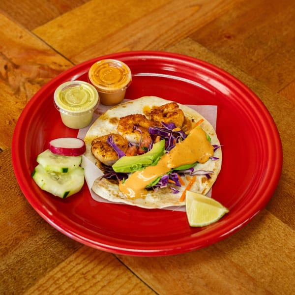 Shrimp taco with avocado, purple cabbage, and orange sauce on a red plate with lime and cucumber.