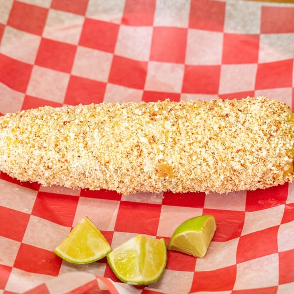Elote, Mexican street corn, covered in cheese and spices, with lime wedges on red checkered paper.