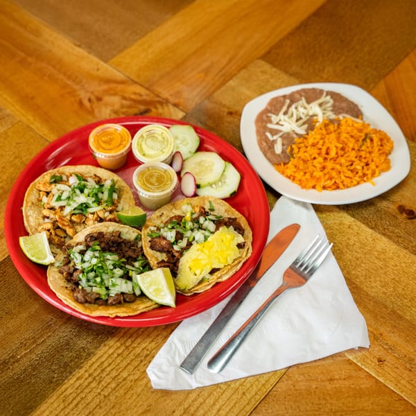 A red plate of Carnales Taqueria tacos with various fillings, lime, sauces, and a side of rice and beans.