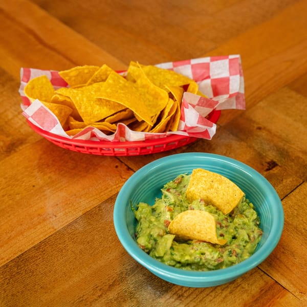 A red basket of tortilla chips and a blue bowl of guacamole with chips on a wooden table.