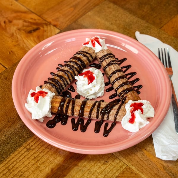 Three churros drizzled with chocolate sauce and topped with whipped cream and strawberry syrup on a pink plate.