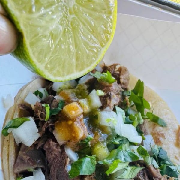 A hand squeezes a fresh lime over a Carnales Taqueria taco filled with meat, onions, and cilantro.