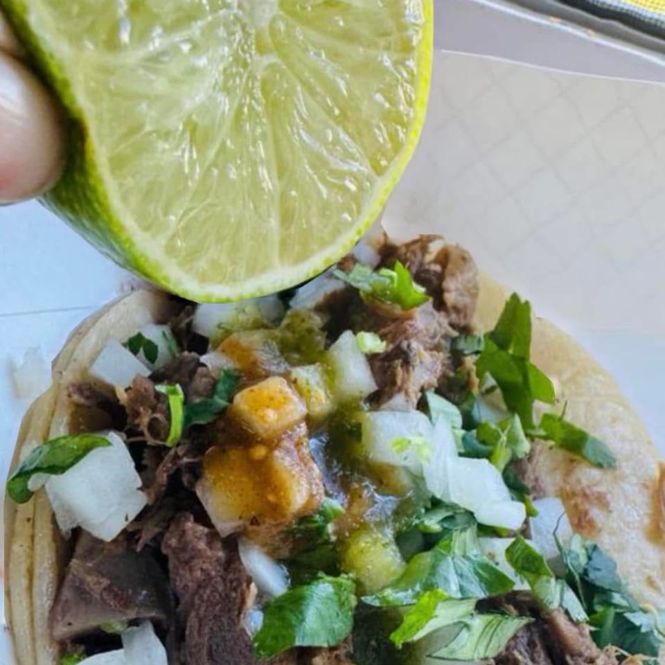 A hand squeezes a fresh lime over a Carnales Taqueria taco filled with meat, onions, and cilantro.