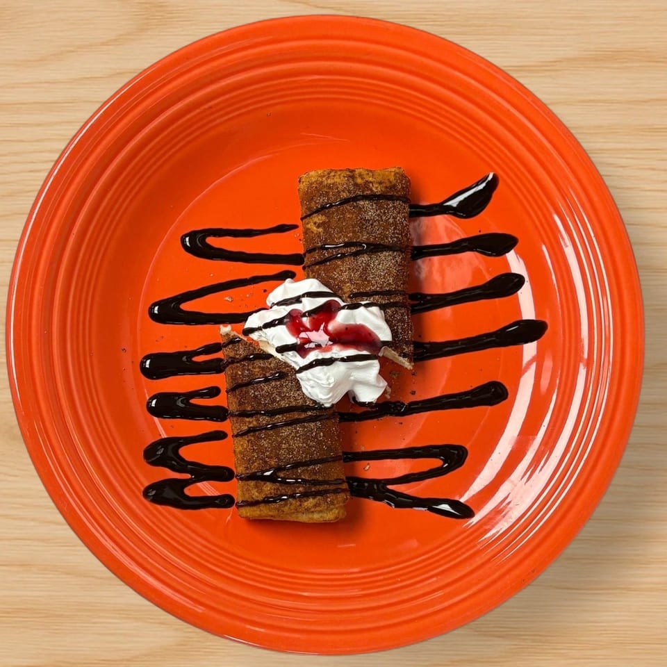 Two churros with whipped cream, strawberry sauce, and chocolate drizzle on an orange plate.