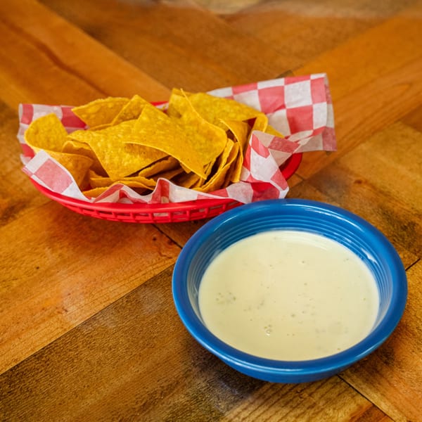 A red basket of tortilla chips and a blue bowl of queso dip on a wooden table.