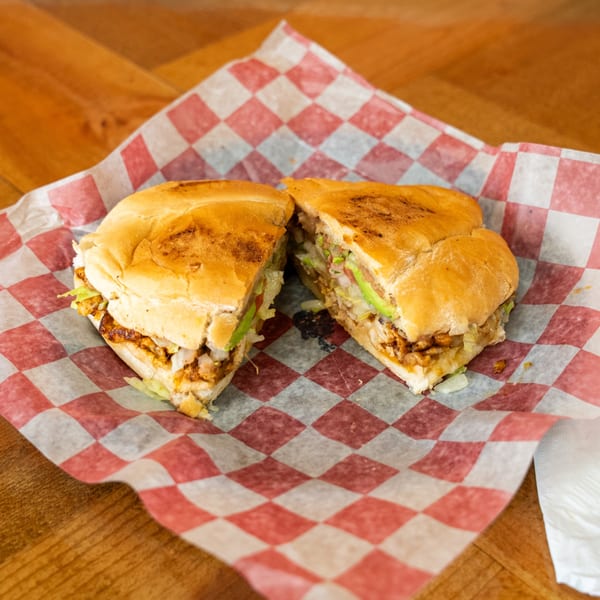 A Carnales Taqueria Mexican torta, cut in half, filled with meat, lettuce, and avocado.