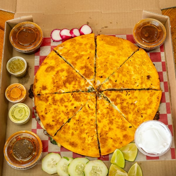 Large, sliced quesadilla with various sauces, radishes, cucumbers, and limes in a takeout box.