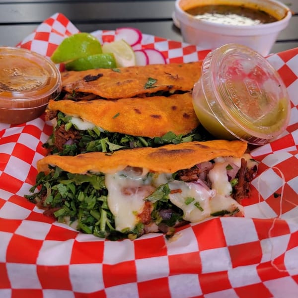 Three Carnales Taqueria birria tacos with melted cheese, cilantro, onions, lime, and sauces.