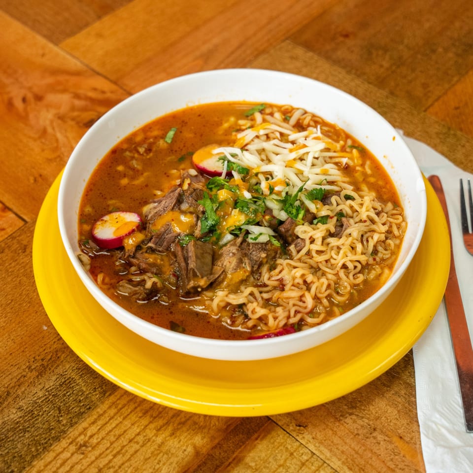 A bowl of Carnales Taqueria's birria ramen with beef, noodles, cheese, cilantro, and radishes.
