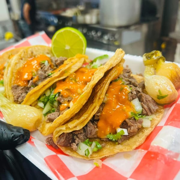 Three Carnales Taqueria tacos with meat, onions, cilantro, and orange sauce, served with lime and grilled onions.
