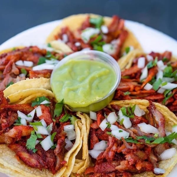 A plate of Carnales Taqueria's al pastor tacos with onions, cilantro, and a side of green salsa.