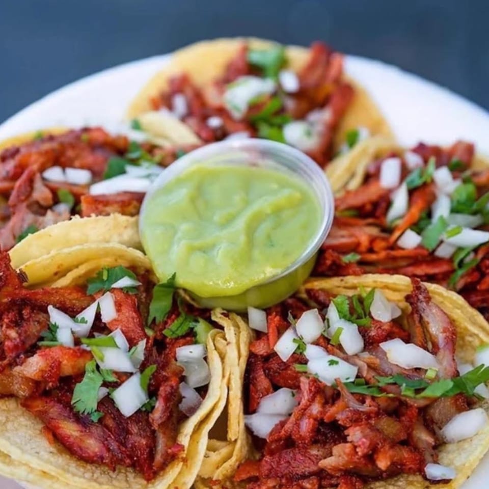 A plate of Carnales Taqueria's al pastor tacos with onions, cilantro, and a side of green salsa.